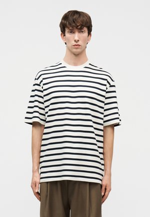 Νεαρός άνδρας που φοράει λευκό T-shirt με κοντά μανίκια και μαύρες οριζόντιες ρίγες και καφέ χαλαρά παντελόνια, στέκεται μπροστά σε απλό φόντο.