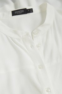 Balta pogām aizsprādzējamā krekla ar apaļu kakla izgriezumu, redzamām pogām un melnu birku ar uzrakstu "SOAKED IN LUXURY M MADE IN CN" apkakles iekšpusē.