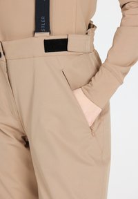 Pantaloni beige con una texture liscia, tasche di dimensioni adeguate e bretelle regolabili con una tracolla nera. Include un dettaglio della tasca con cerniera.
