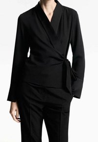 Blouse portefeuille noire en tissu lisse, dotée de manches longues, d'un décolleté en V et d'un détail noué à la taille pour une silhouette ajustée.