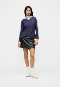 Γυναίκα με μακριά μαύρα μαλλιά, που φοράει μπλε πουλόβερ με γιακά, μαύρη δερμάτινη φούστα, λευκές κάλτσες και λευκά αθλητικά παπούτσια, στέκεται μπροστά σε απλό φόντο.