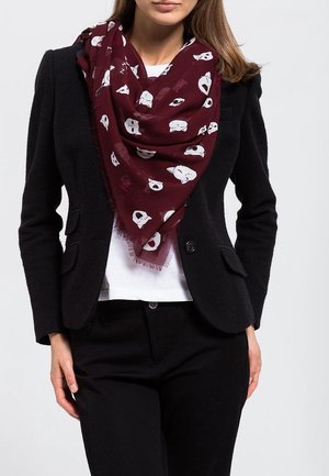 Femme portant un blazer noir et un pantalon assorti avec une chemise blanche, écharpe bordeaux ornée de motifs de têtes d'animaux blanches.