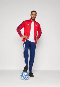 Rdeča Adidas trenirka z modrimi črtami, bela majica, modre trenirke hlače z belimi črtami in črni čevlji; stoji na nogometni žogi.