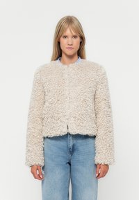 Giacca cropped beige chiaro realizzata in un tessuto fluffy e strutturato, con colletto rotondo e maniche lunghe, abbinata a jeans di denim blu.