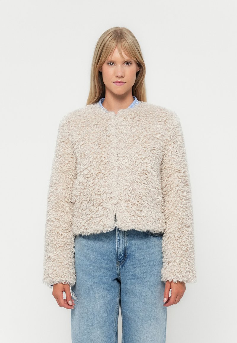 Giacca cropped beige chiaro realizzata in un tessuto fluffy e strutturato, con colletto rotondo e maniche lunghe, abbinata a jeans di denim blu.