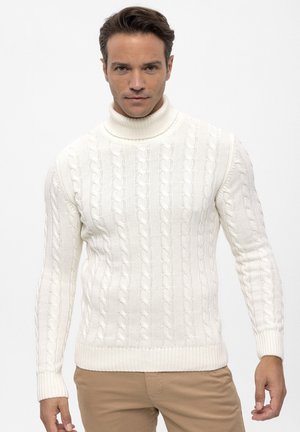 Homme portant un pull à col roulé torsadé blanc et un pantalon beige sur un fond clair uni.