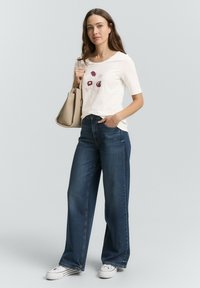 Witte t-shirt met korte mouwen en bloemenontwerp, gecombineerd met donkerblauwe wijde jeans en witte sneakers, vastgehouden met een beige handtas.