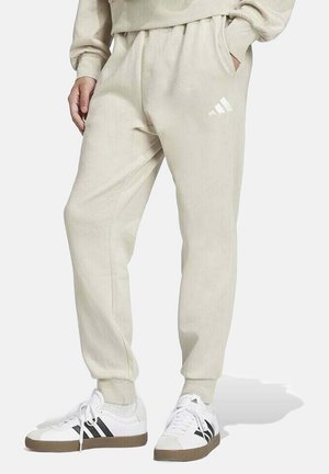 Pantaloni jogger beige realizzati in materiale morbido, con vita elastica, tasche laterali e un logo Adidas bianco sulla coscia sinistra.