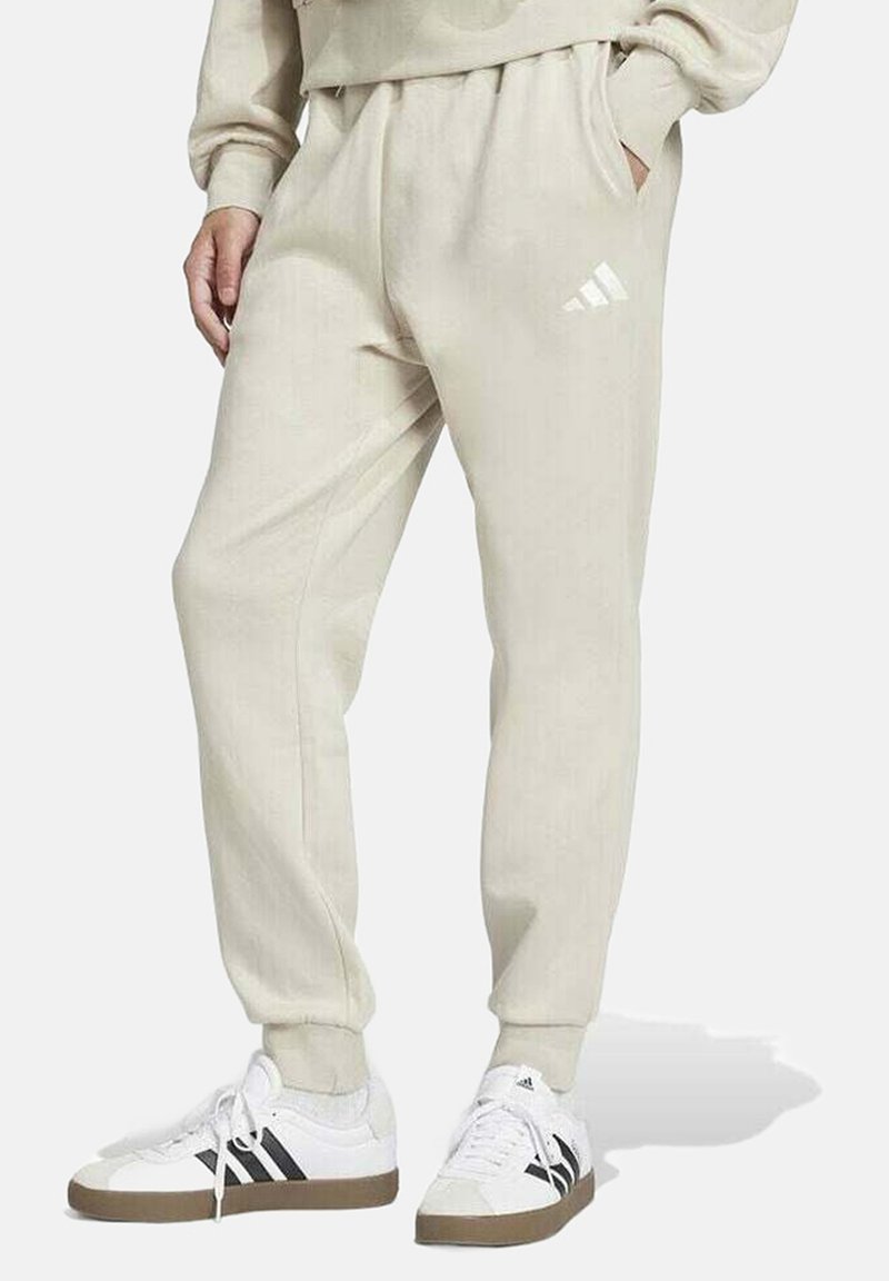 Pantaloni jogger beige realizzati in materiale morbido, con vita elastica, tasche laterali e un logo Adidas bianco sulla coscia sinistra.