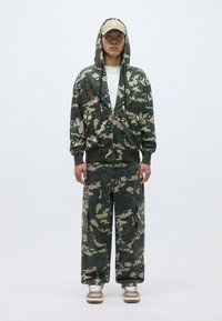 Kamuflāžas zip-пulovers un kravas bikses zaļos un brūnos toņos, izgatavotas no mīkstas auduma, ar lielām kabatām un atbilstošu cepuri.