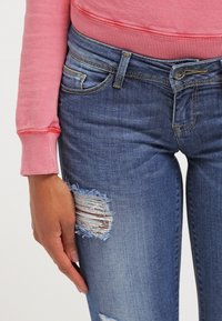 Jeans de mezclilla azul ajustados con detalles desgastados en las rodillas, con una cintura de altura media y una sudadera rosa en contraste con puños acanalados.