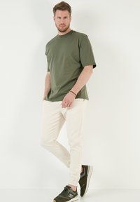 Olivově zelené tričko s krátkým rukávem, krémové joggingové kalhoty a tmavé tenisky s oranžovými akcenty; model stojí na bílém pozadí.