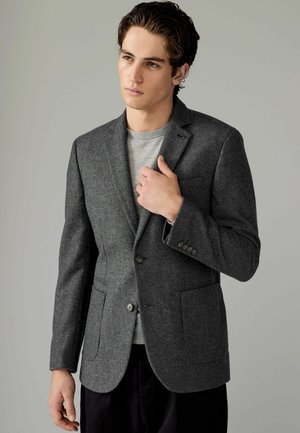 Grauer Fischgrät-Blazer mit zwei Knöpfen, Reverskragen und seitlichen Taschen. Über einem hellgrauen Hemd getragen. Strukturierter Wollstoff.