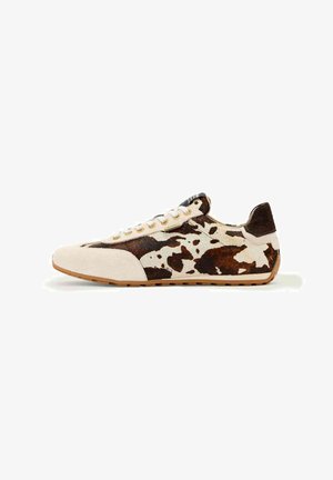Lavprofil sneakers med hvidt og brunt ko-mønstret print, beige ruskindsdetaljer, hvide snørebånd og sål i gummikorn, vist fra siden.