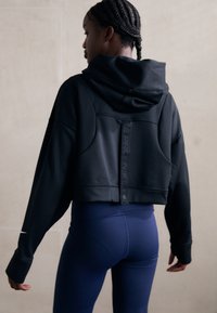 Svart avkortet hettegenser med hette og fremtredende sømdetaljer, med en reflekterende Nike-merkingstropp bak. Kombinert med blå leggings.
