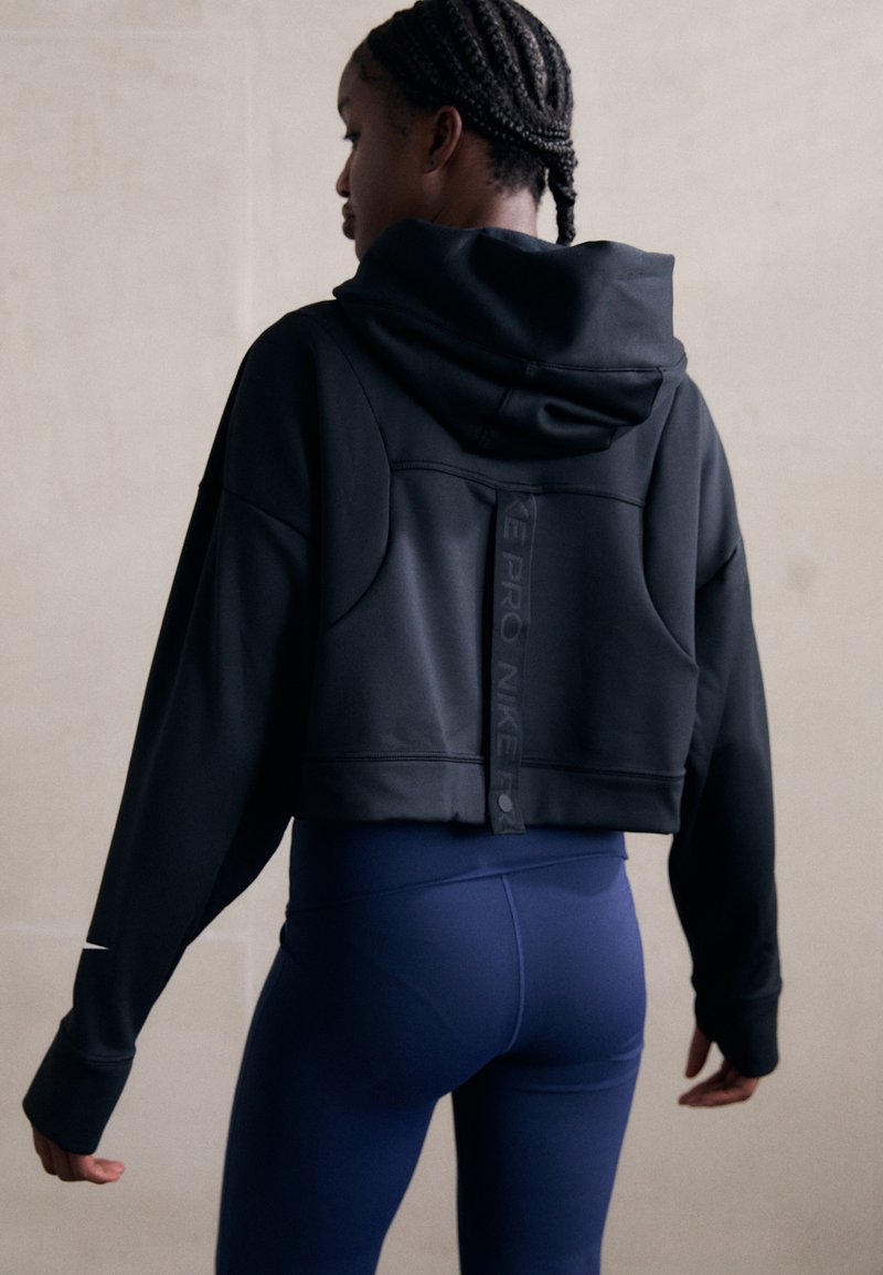 Sweatshirt noir court avec capuche et détails de couture remarquables, doté d'une sangle de marque réfléchissante Nike à l'arrière. Associé à des leggings bleus.