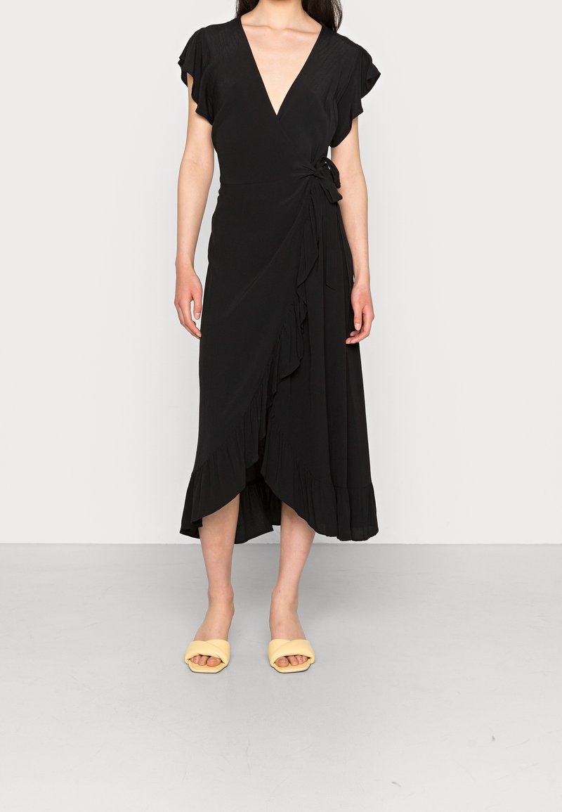 Robe portefeuille noire avec manches évasées, un col en V profond et un ourlet volanté. Assortie à des sandales slides jaunes sur une surface lisse gris clair.