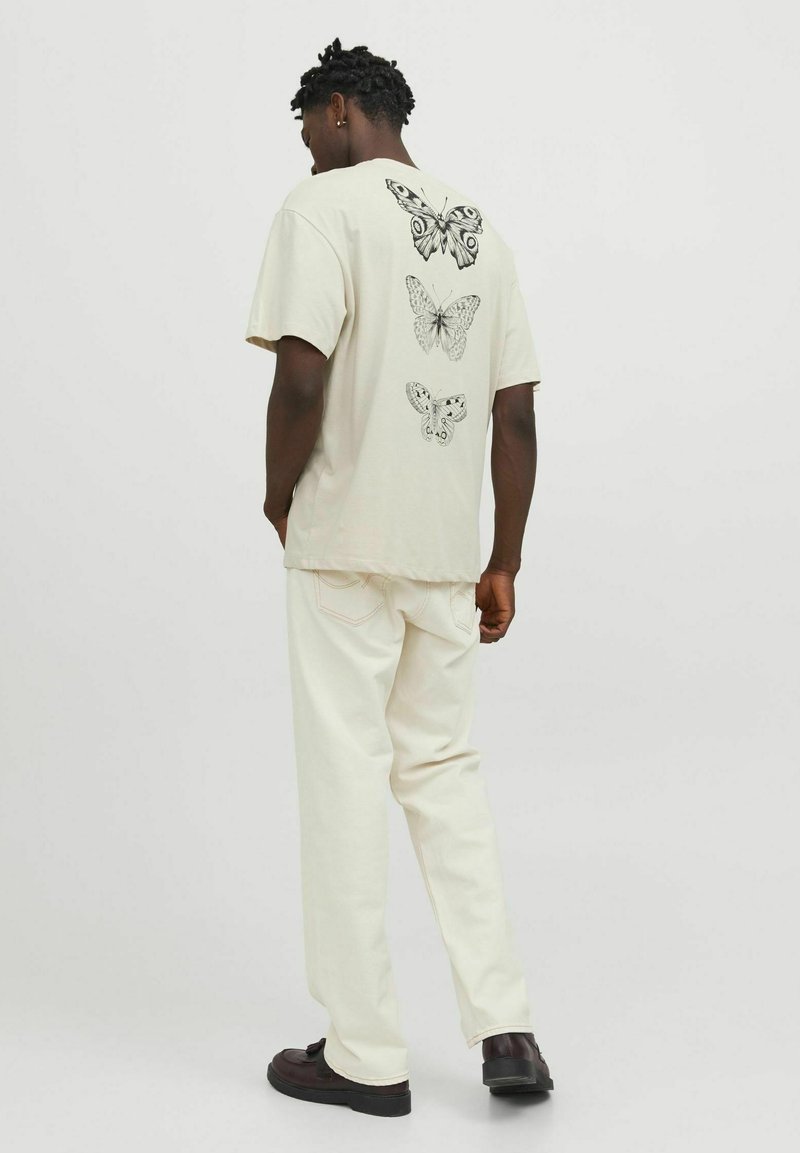 Cream short-sleeve t-shirt with black butterfly illustrations on the back, paired with light-colored pants and dark shoes.