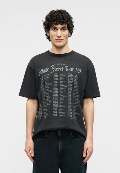 Νεαρός με κατσαρά μαλλιά που φοράει μια μαύρη φαρδιά μπλούζα με καταχωρημένες ημερομηνίες και πόλεις περιοδείας από το 1999 «Infinite Secret Tour».