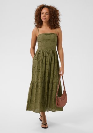 Femme aux cheveux bouclés portant une robe midi ajourée vert olive, des sandales noires et tenant un sac à bandoulière marron, debout sur fond blanc.
