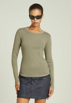 Haut à manches longues vert olive avec une texture côtelée, col rond et ourlet incurvé, associé à une jupe en denim foncé et des lunettes de soleil noires élégantes.