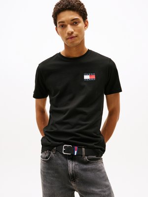 Sort bomulds t-shirt med et lille farverigt logo patch, parret med mørke denim jeans og et sort bælte med røde, hvide og blå detaljer.