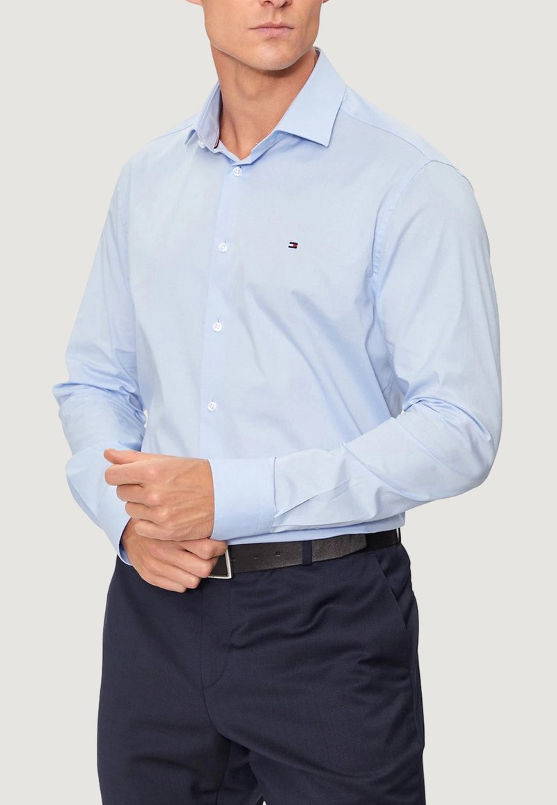 Camicia azzurra chiaro con bottoni e maniche lunghe, realizzata in tessuto liscio. Presenta un piccolo logo sul petto e un colletto aperto. Abbinata a pantaloni scuri.