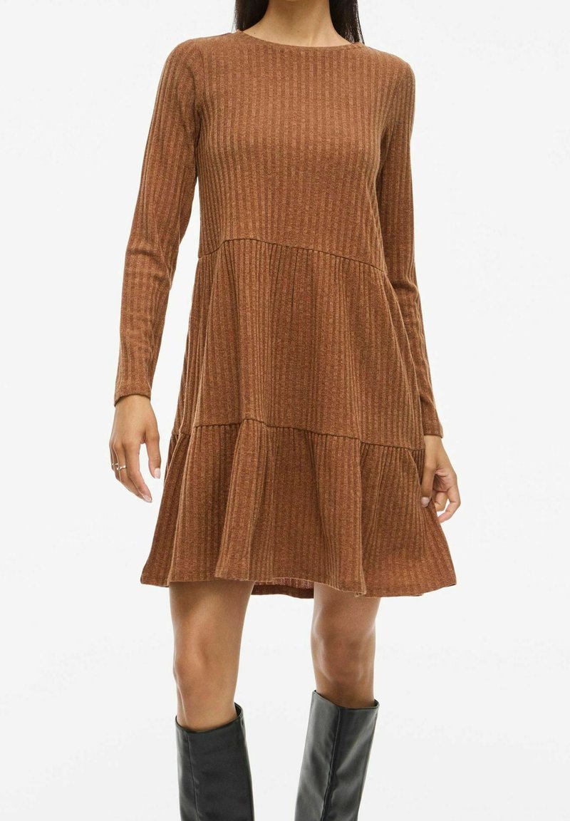 Femme portant une robe marron côtelée à manches longues à volants, associée à des bottes noires mi-mollet, sur fond blanc uni.