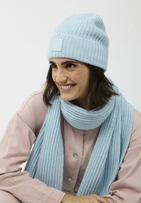 Lichtblauwe gebreide beanie met ribbels en logopatch, gecombineerd met een bijpassende lichtblauwe sjaal met een vergelijkbaar breipatroon.
