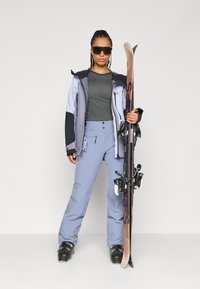Îmbrăcăminte de schi cu un top gri, pantaloni de culoare albastră deschisă și o jachetă bicoloră cu accente negre. Modelul ține schiuri și poartă ochelari de soare închis.