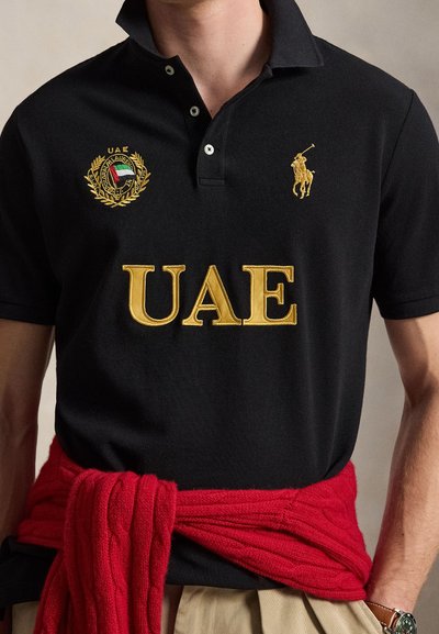 Μαύρο πόλο με χρυσό κείμενο "UAE", έμβλημα με θέμα τα ΗΑΕ και λογότυπο παίκτη πόλο, φορεμένο με κόκκινο πουλόβερ δεμένο γύρω από τη μέση και μπεζ παντελόνια.