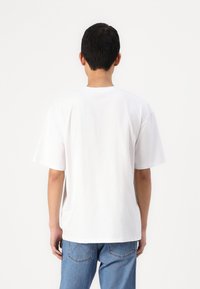 T-shirt alb din bumbac, cu mâneci scurte, croială lejeră și decolteu rotund. O pereche de blugi albastru deschis cu buzunare posterioare vizibile.