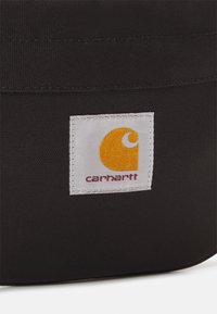 Czarna torba z materiału z białą etykietą z żółtym stylizowanym logo "C" oraz napisem "carhartt". Gładka tekstura, prostokątny kształt, solidne szwy.