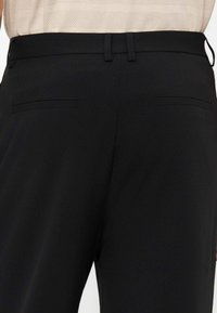 Pantalon noir sur mesure avec passants de ceinture et deux poches arrière horizontales, porté avec un top crème texturé rentré.