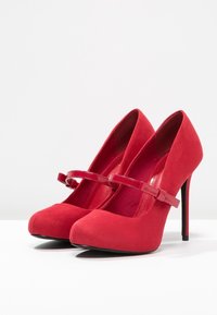 Red suede high heels with a pointed toe, a glossy strap across the vamp, and a thin stiletto heel. Smooth texture and vibrant color.