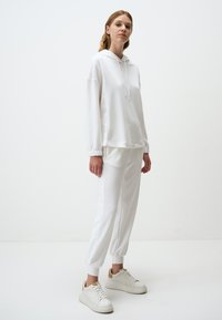 Witte hoodie met lange mouwen en verstelbare koord, gecombineerd met bijpassende tapered broek. Witte sneakers met beige accenten maken de look compleet.