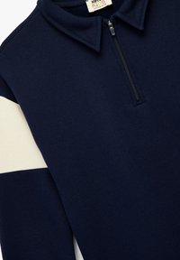 Granatowy pulower z suwakiem do połowy z kołnierzykiem, z białym paskiem na lewym rękawie. Miękka tkanina o gładkiej fakturze i minimalistycznym designie.