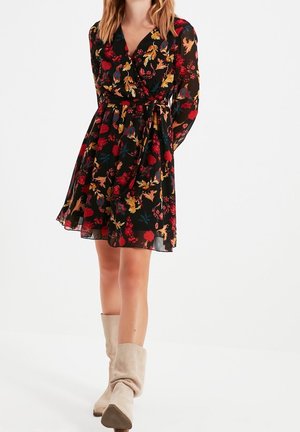Vrouw draagt een zwarte bloemenprint wikkeljurk met lange mouwen en beige slappe kniehoge laarzen, staande tegen een witte achtergrond.