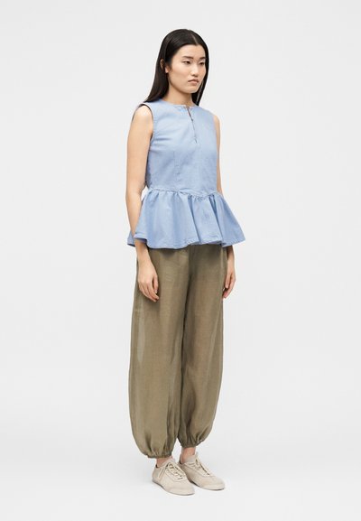 Femme portant un top bleu clair sans manches avec basque, un pantalon ample vert olive avec des poignets élastiques et des baskets beiges, debout devant un fond uni.