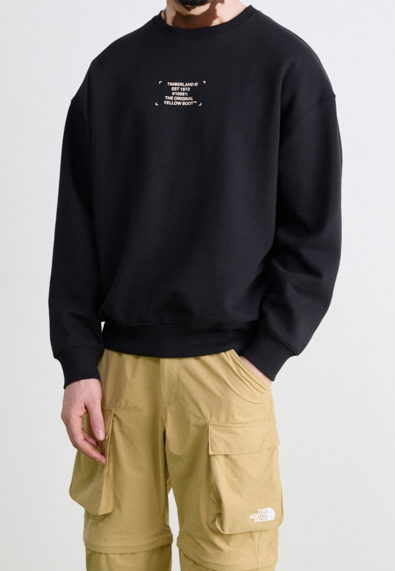 Homme portant un sweat Timberland noir avec texte et un pantalon cargo beige The North Face avec logos sur les poches, mains dans les poches, sur fond uni.