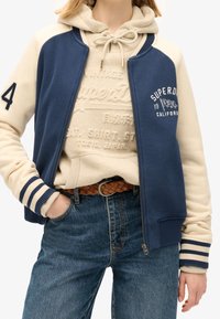 Granatowo-kremowa kurtka sportowa z prążkowanymi mankietami, zamkiem na przedzie i haftowanym logo. Na wierzchu beżowa bluza z kapturem z nadrukiem. Jeansy.