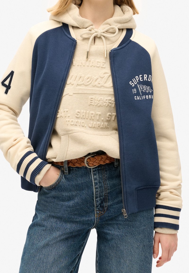 Granatowo-kremowa kurtka sportowa z prążkowanymi mankietami, zamkiem na przedzie i haftowanym logo. Na wierzchu beżowa bluza z kapturem z nadrukiem. Jeansy.