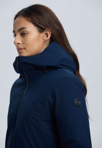 Marineblauwe jas met een hoge kraag, ritssluiting en verstelbare capuchon. Opvallende logopatch op de mouw en een gladde textuur.