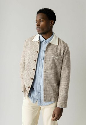 Mand iført beige stribet knapjakke over lyseblå skjorte og cremebukser, ser tankefuldt til siden mod en ensfarvet baggrund.