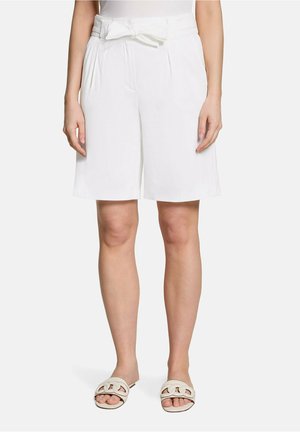Femme portant un short blanc taille haute avec ceinture nouée à l'avant et des sandales blanches plates à bout ouvert, debout devant un fond uni.