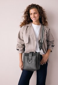 Black leather crossbody bag with a front flap, silver hardware, and a patterned adjustable strap. Worn with a beige jacket and white top.
