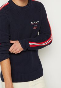 Marineblauer Strickpullover mit gerippten Bündchen und Saum. Kontrastierende rot-weiße und blaue gestreifte Details entlang der Ärmel. Gesticktes Logo auf der Brust.