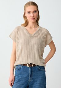 Femme aux cheveux châtain clair portant un top beige côtelé à manches courtes et col en V, rentré dans un jean bleu avec une ceinture marron, debout devant un fond uni.