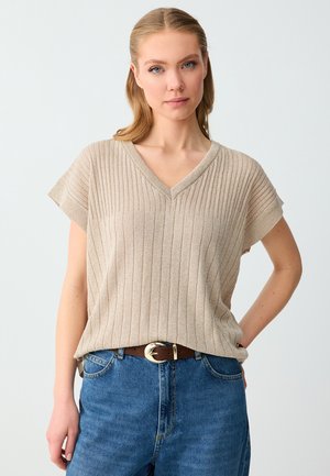 Femme aux cheveux châtain clair portant un top beige côtelé à manches courtes et col en V, rentré dans un jean bleu avec une ceinture marron, debout devant un fond uni.