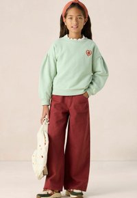 Sweatshirt vert menthe avec des manches bouffantes et une broderie en forme de cœur, associé à un pantalon bordeaux large. Un sac beige avec des accents à motifs.